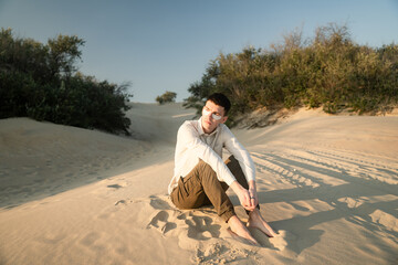 a young guy in a beige shirt and pants sits on the hot sand, white patches on his face