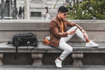 Obraz premium Happy young handsome man wearing spring clothes sitting outdoor in park talking and texting on cellphone