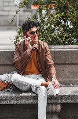 Obraz premium Happy young handsome man wearing spring clothes sitting outdoor in park talking and texting on cellphone