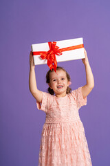 little cute girl with gift box with red ribbon on purple background. birthday of small child. a happy child holds a gift in his hands. a birthday present. concept of celebrating, giving and receiving
