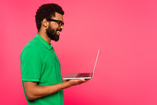 Side View Of Happy African American Man In Glasses And Green Polo Shirt Holding Laptop Isolated On Pink