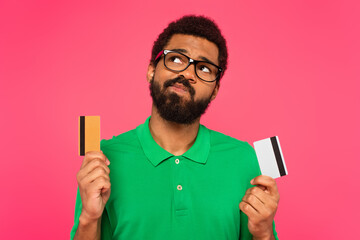 confused african american man in glasses holding credit cards isolated on pink - obrazy, fototapety, plakaty