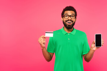 amazed african american man in glasses holding smartphone and credit card isolated on pink