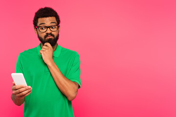 pensive african american man in green t-shirt using smartphone isolated on pink