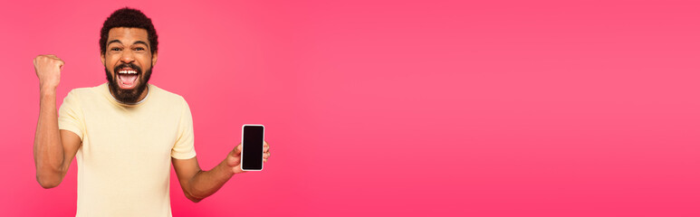 excited african american man holding smartphone with blank screen isolated on pink, banner