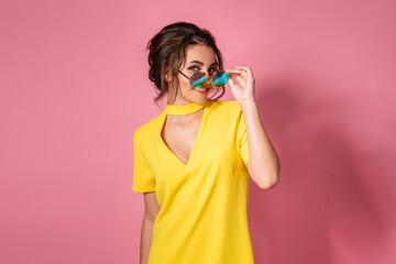 Beautiful girl in yellow dress wearing sunglasses posing, smiling on pink background in studio. 
