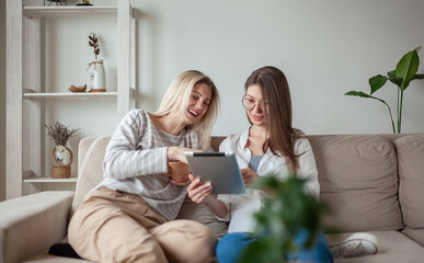 Two young women friends using tablet together, sitting on the couth