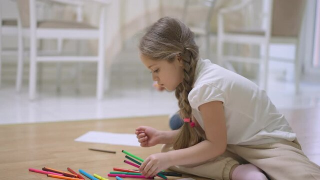 Side view of beautiful Caucasian little girl with pigtails choosing colorful markers with baby sister drawing at background. Creative siblings enjoying hobby indoors at home. Childhood lifestyle