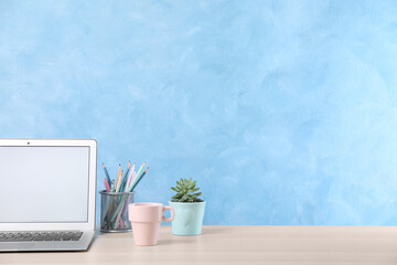 Modern laptop with blank screen, cup and office supplies on white table near light blue wall, space...