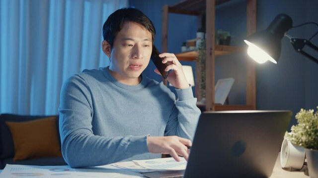 Young Asia businessman use smartphone call meeting agenda assignment paperwork with colleague look at laptop computer in living room at home overtime at night, Work from home corona pandemic concept.