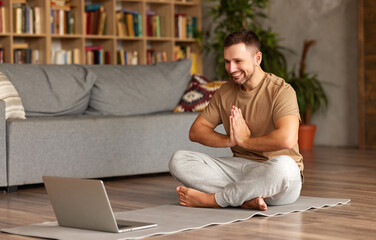 Fototapeta premium Young cheerful man in casual wear sitting in lotus pose in front of laptop at home
