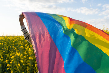 Unrecognizable man holding rainbow flag in yellow flowers field at sunset.Summer celebration...