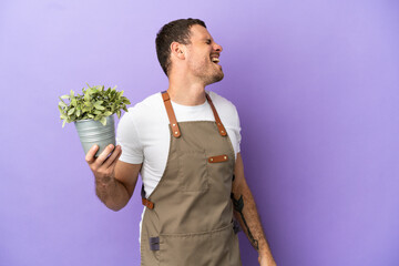 Brazilian Gardener man holding a plant over isolated purple background laughing in lateral position