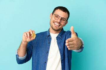 Brazilian man holding a Bitcoin over isolated blue background shaking hands for closing a good deal