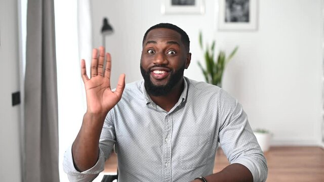 Cheerful and enthusiastic African-American guy wearing smart casual shirt waving hello into the camera, greeting online interlocutor, says hi into webcam. Virtual meeting concept
