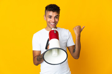 Young brazilian man isolated on yellow background shouting through a megaphone and pointing side