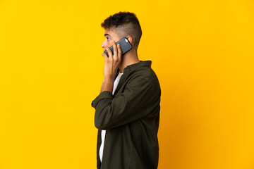 Young brazilian man isolated on yellow background keeping a conversation with the mobile phone with someone