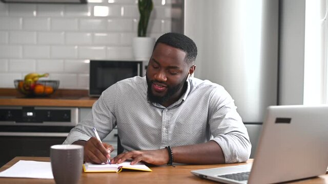 Hard-working African American man sitting at the table at home in the modern kitchen and making notes, trying to remember smth, using the laptop for distance video communication, studying, working,