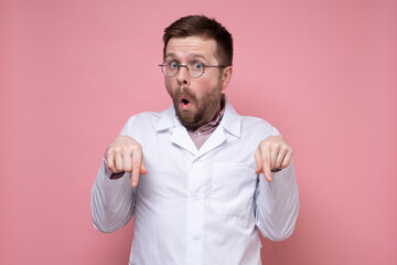 Amazed, shocked young doctor points down with index fingers and looks with large eyes with an open mouth. Pink background.