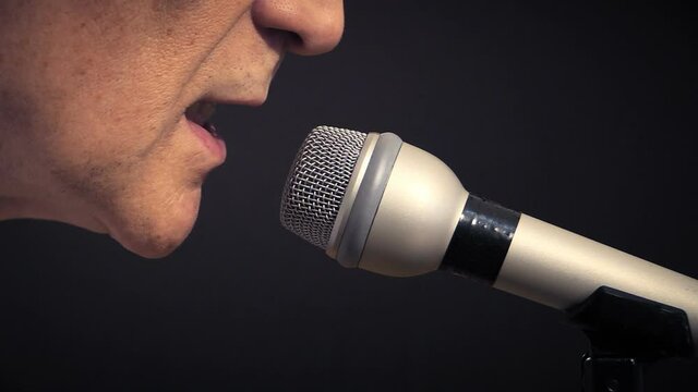 A man in a close up shot, talking into a microphone on a black background