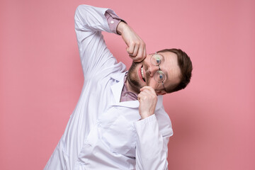 Funny doctor makes a smile by holding mouth with hands, he does not lose heart and entertains patients. Pink background.