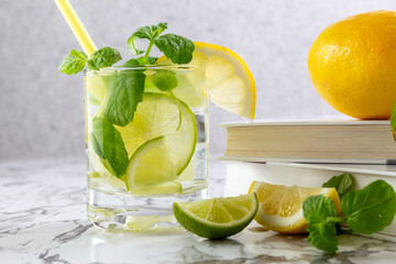 Citrus lemonade with ice in a glass decorated with lemon, lime and mint slices and books on a marble table. Fresh summer drink. Detox. Healthy food rich in vitamins and antioxidants. Copy space