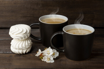 Fototapeta na wymiar hot coffee and marshmallows on a dark woody background. two black cups of coffee with steam and marshmallows decorated with white flowers.