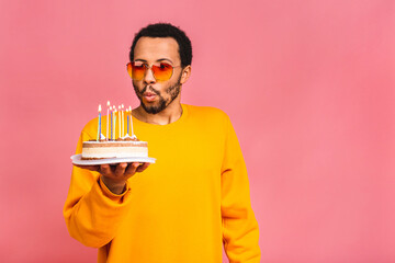 Joyful young african american man blowing candles on a birthday cake isolated on pink background.