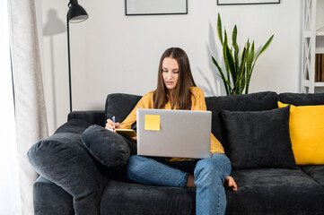 Focused woman freelancer researching business tasks, taking a notes with a laptop at home. A female...