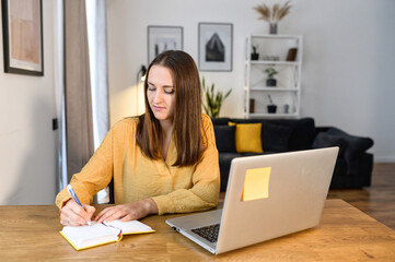 Online training. Female student watching a lecture, webinar using a laptop, sits at the table and writes with a pen in a notepad. A young woman takes a note watching online training