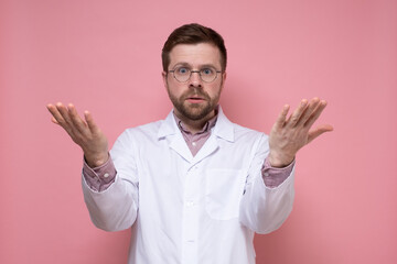 Young bearded doctor in round glasses and a white coat is in perplexity, he throws hands up and looks displeased. Pink background.