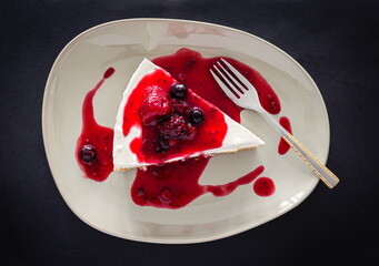 Slice of plain homemade cheesecake with forest fruits sauce on beige plate with a small fork. Black background table. Top view.
