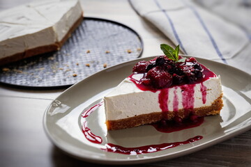 Slice of plain homemade cheesecake with forest fruits sauce decorated with mint leaf on beige plate with a small fork, glass of red fruit juice . Black background table, flower pot. Top view.