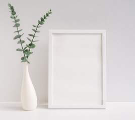 Mock up white wooden poster frame decor with  Eucalyptus  leaves  in modern white vase on white table and wall background