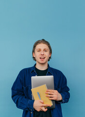 Obraz premium Vertical portrait of a handsome young man in a shirt with a laptop and a notebook in his hands isolated on a blue background, looking at the camera and smiling.