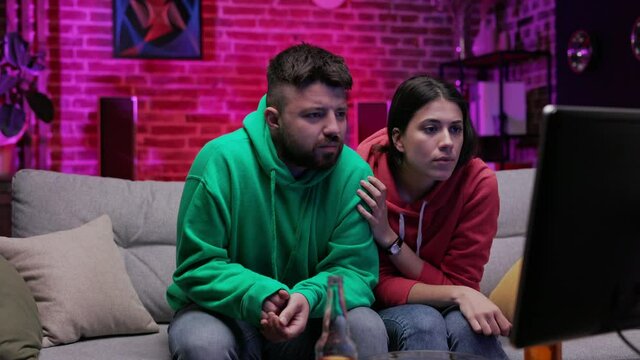 Caucasian young girlfriend and boyfriend watching horror movie on the TV in the living room at night. Girl frightened by scarry moment and hiding at her boyfriend shoulder. Concept of relationship.