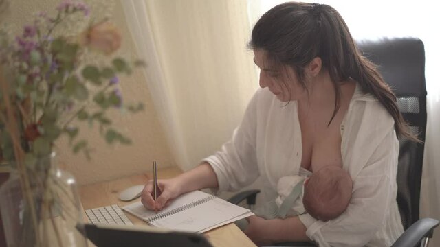 Mom breastfeeding baby and writing in copybook in house