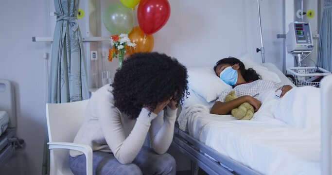 Stressed african american mother sitting besides her her daughter lying on bed at hospital
