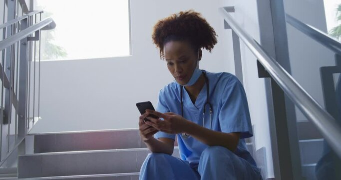 Mixed race female doctor wearing face mask sitting on stairs in hospital using smartphone