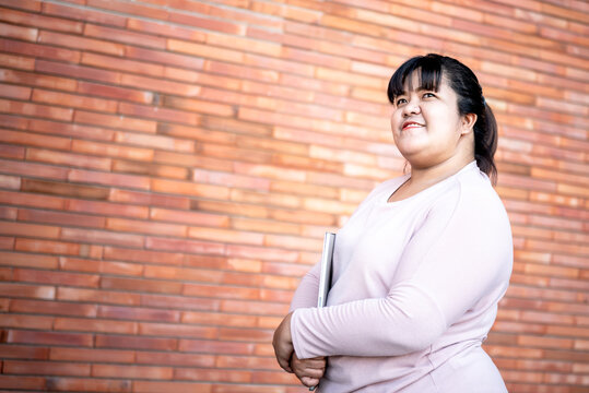 Portrait images of Asian attractive fat woman standing holding computer notebook and looking up, with wall background, to people and adult education concept.