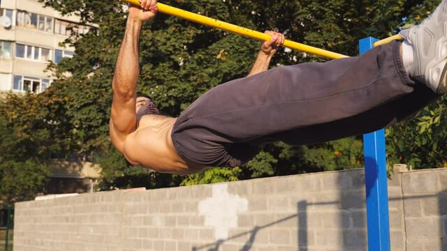 Close up to young muscular athlete showing some gymnastics stunts on horizontal bar outdoor. Strong sporty man performing static exercises during workout at sports ground. Sportive lifestyle concept