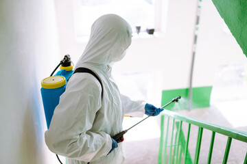 Man wearing protective suit disinfecting the entrance of a residential building with spray...