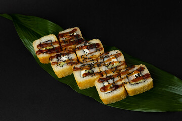 Tempura is a deep-fried sushi roll with prawns, cucumber and cream cheese inside, dressed with sauce and sesame seeds. Sushi roll on a banana leaf, on a black background. Japanese sushi food.