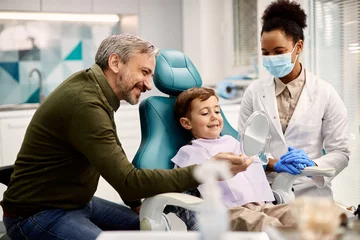Happy boy looking at his teeth in a mirror while being with his father at dentist's office.