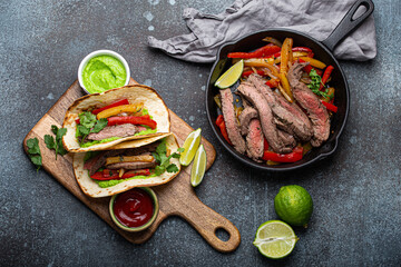 Process of making traditional Mexican dish Beef fajitas tacos served on wooden cutting board with tomato salsa and guacamole on rustic stone background from above, American Mexican food 