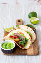 Traditional Mexican dish Beef fajitas tacos served on wooden cutting board with tomato salsa and guacamole on rustic white wooden background from angle view, American Mexican food 