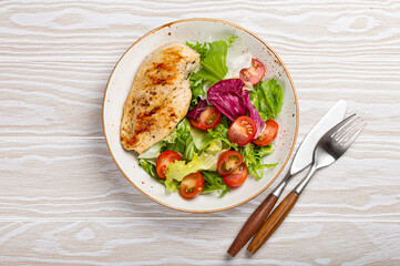 Grilled chicken breast fillet with vegetables salad of fresh tomatoes and salad leaves on white plate on wooden background from above, healthy dinner or diet meal 
