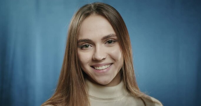 Portrait of beautiful teenager smiling, posing in fashion studio on blue background. Close-up of young cute woman, model touching her hair. 