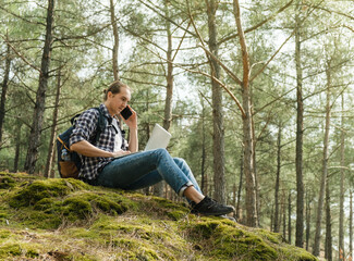 Naklejka na ściany i meble Young man sitting on ground at forest talking on smartphone and working on laptop
