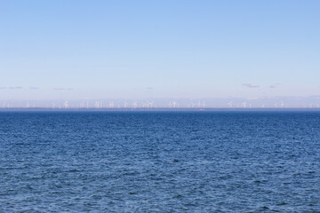 Obraz premium A view from open water of a wind turbine farm on the far shore as seen from a ferry - light hazy blue sky over dark blue water